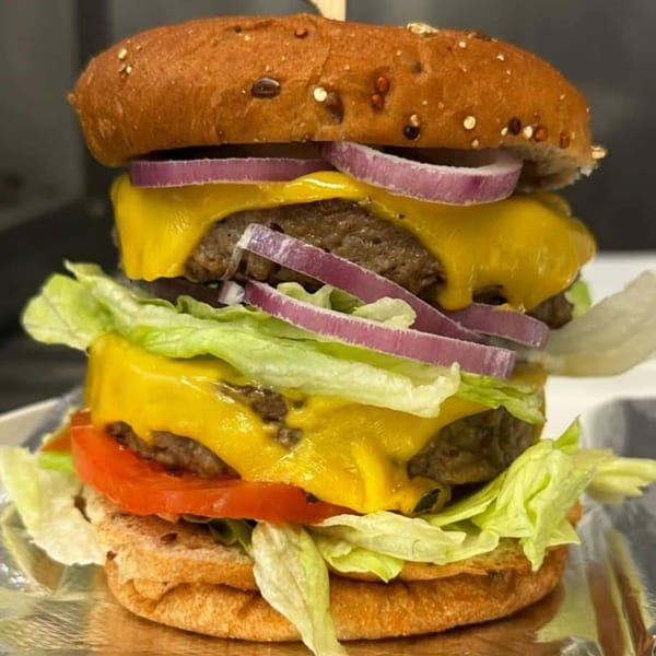 A towering vegan double cheeseburger with lettuce, tomato, and red onion on a pretzel bun.