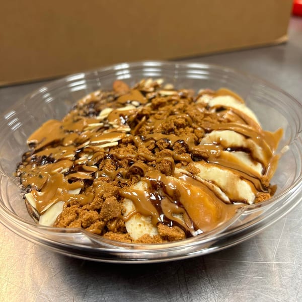 A vegan smoothie bowl with banana slices, cookie crumbles, and caramel drizzle in a clear plastic container.