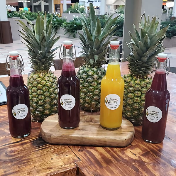 Five Essential Smoothies Cafe juices in bottles, with four whole pineapples, on a wooden table.
