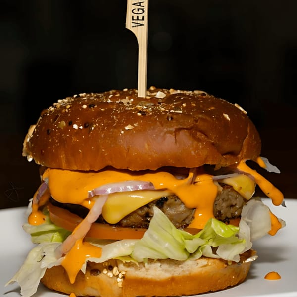 A vegan burger with a patty, melted cheese, lettuce, tomato, red onion, and orange sauce.